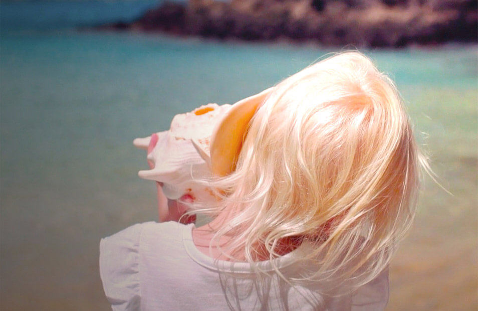 Photo of blond girl holding a seashell to her ear and looking at the ocean